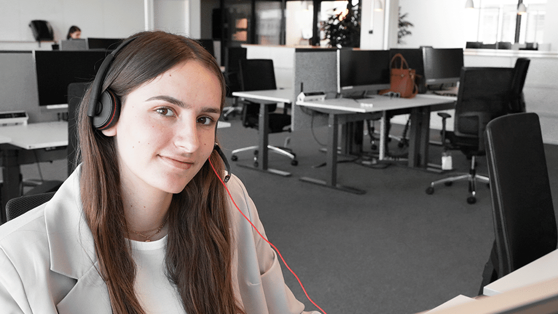 Eine junge Frau mit Headset und hellem Blazer lächelt in die Kamera, während sie an einem Schreibtisch in einem modernen Großraumbüro mit leeren Arbeitsplätzen im Hintergrund sitzt.
