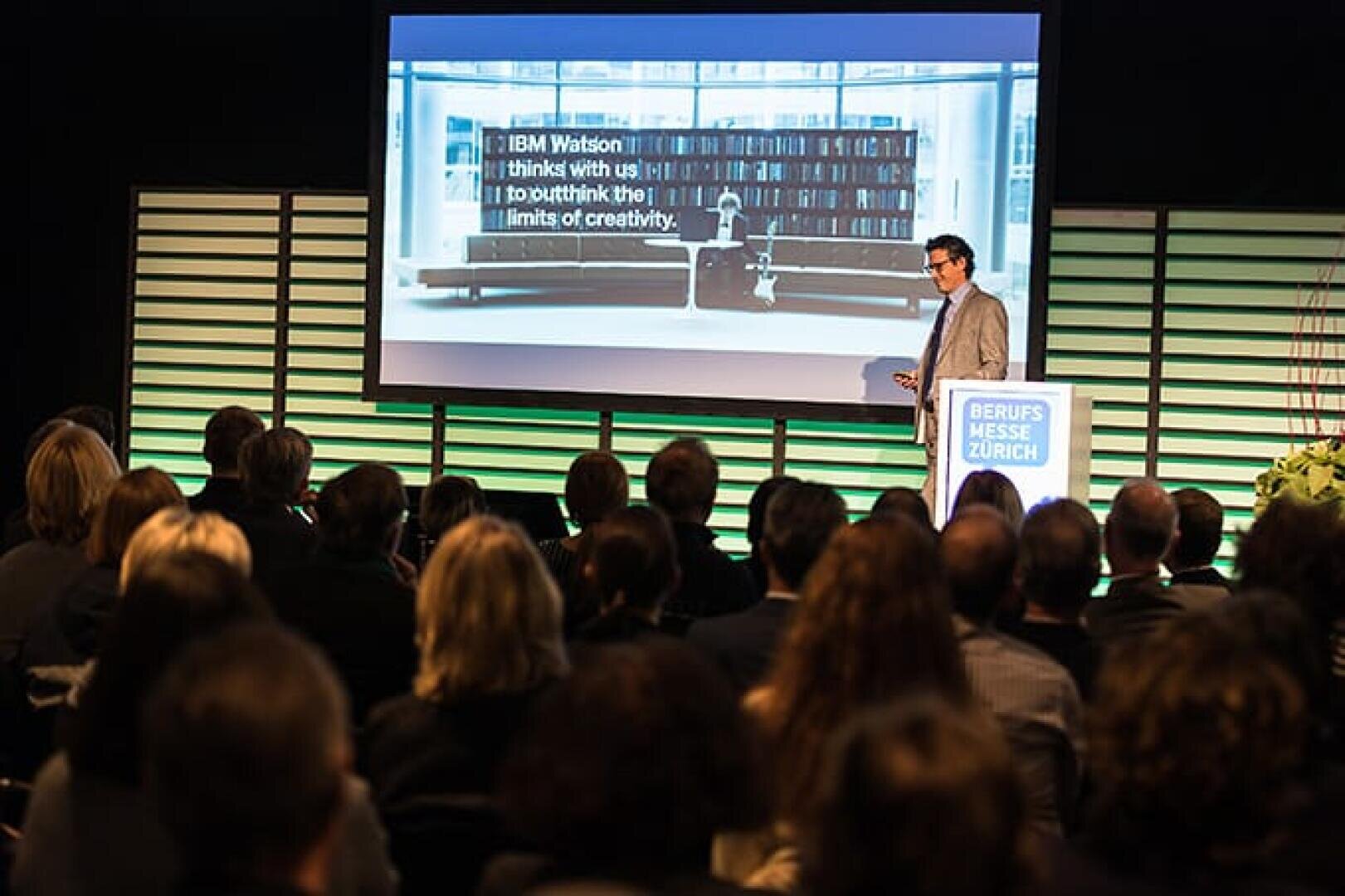 Ein Redner präsentiert dem Publikum an der Beruf Messe Zürich eine Folie über IBM Watson mit dem Zitat "Ich denke mit uns! um die Grenzen der Kreativität zu überschreiten" in einer modernen Konferenzumgebung.