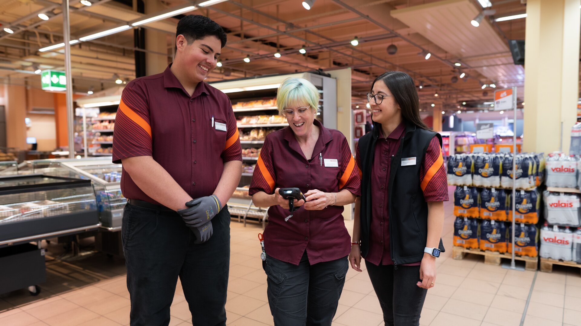Drei Supermarktangestellte in passenden Uniformen stehen zusammen in einem Gang, lächeln und unterhalten sich. Eine Person hält einen Handscanner in der Hand, während die anderen darauf schauen. Im Hintergrund sind Regale und Produkte zu sehen.
