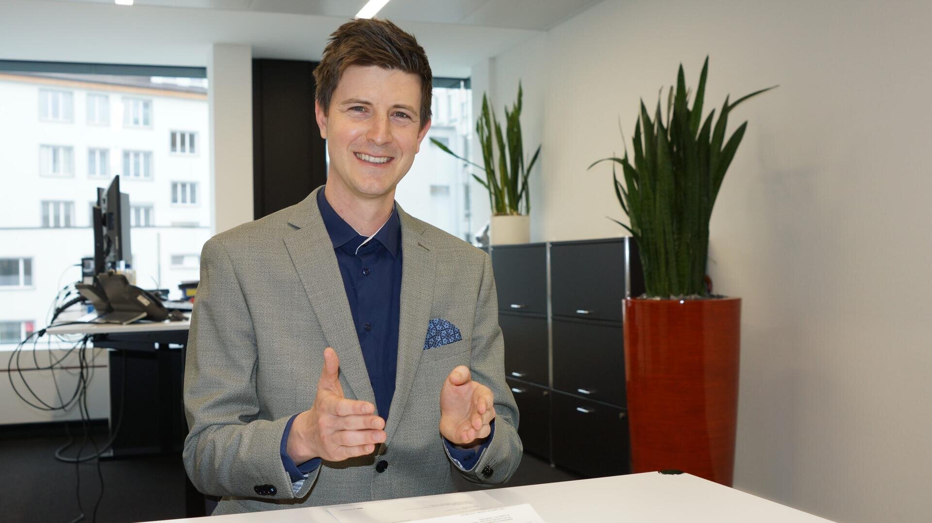 Ein lächelnder Mann in einem grauen Anzug gestikuliert mit den Händen, während er an einem Schreibtisch in einem modernen Büro sitzt. Hinter ihm stehen hohe Grünpflanzen und schwarze Aktenschränke, große Fenster lassen viel Tageslicht herein.