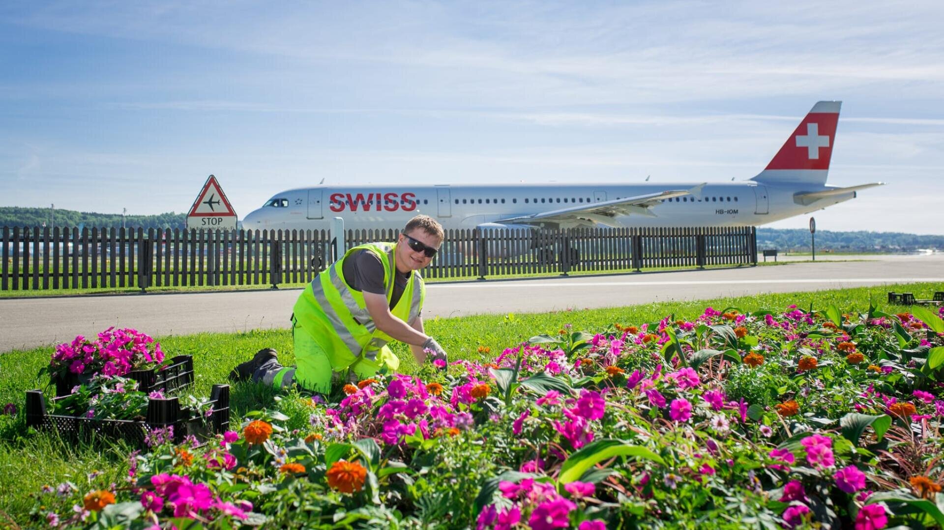 Eine Person in einer neonfarbenen Sicherheitsweste kniet inmitten bunter Blumen und pflegt einen Garten in der Nähe einer Flughafenpiste. Im Hintergrund ist ein Schweizer Flugzeug zu sehen, in der Nähe befinden sich ein Zaun und ein Stoppschild.