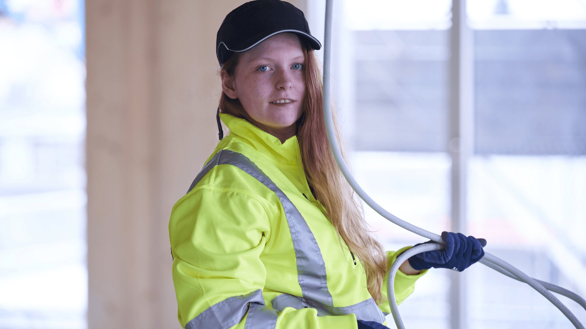Eine Frau mit schwarzer Mütze, gelber Warnweste und Handschuhen hält ein Spiralkabel in einer Baustelle und blickt in die Kamera.