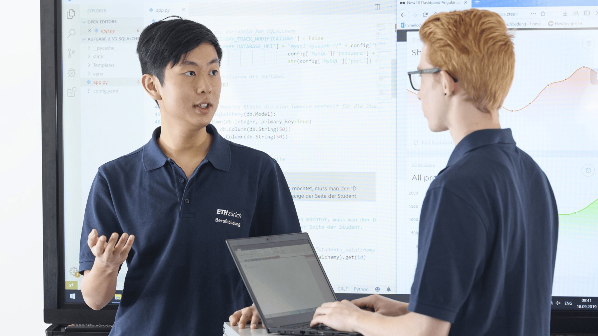 Zwei junge Männer in marineblauen Hemden diskutieren etwas, während einer einen Laptop hält. Sie stehen vor Bildschirmen, auf denen Codes und Datendiagramme angezeigt werden. Beide scheinen konzentriert und in ein Gespräch vertieft zu sein.