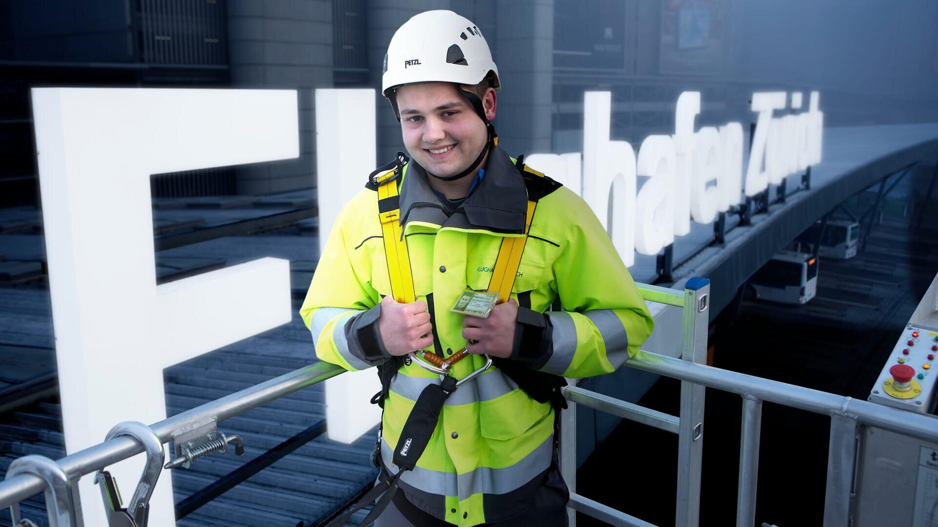 Ein Arbeiter in Warnweste, Helm und Sicherheitsgurt lächelt, während er auf einer Hebebühne vor einer großen weißen Beschilderung an einem Gebäude steht.