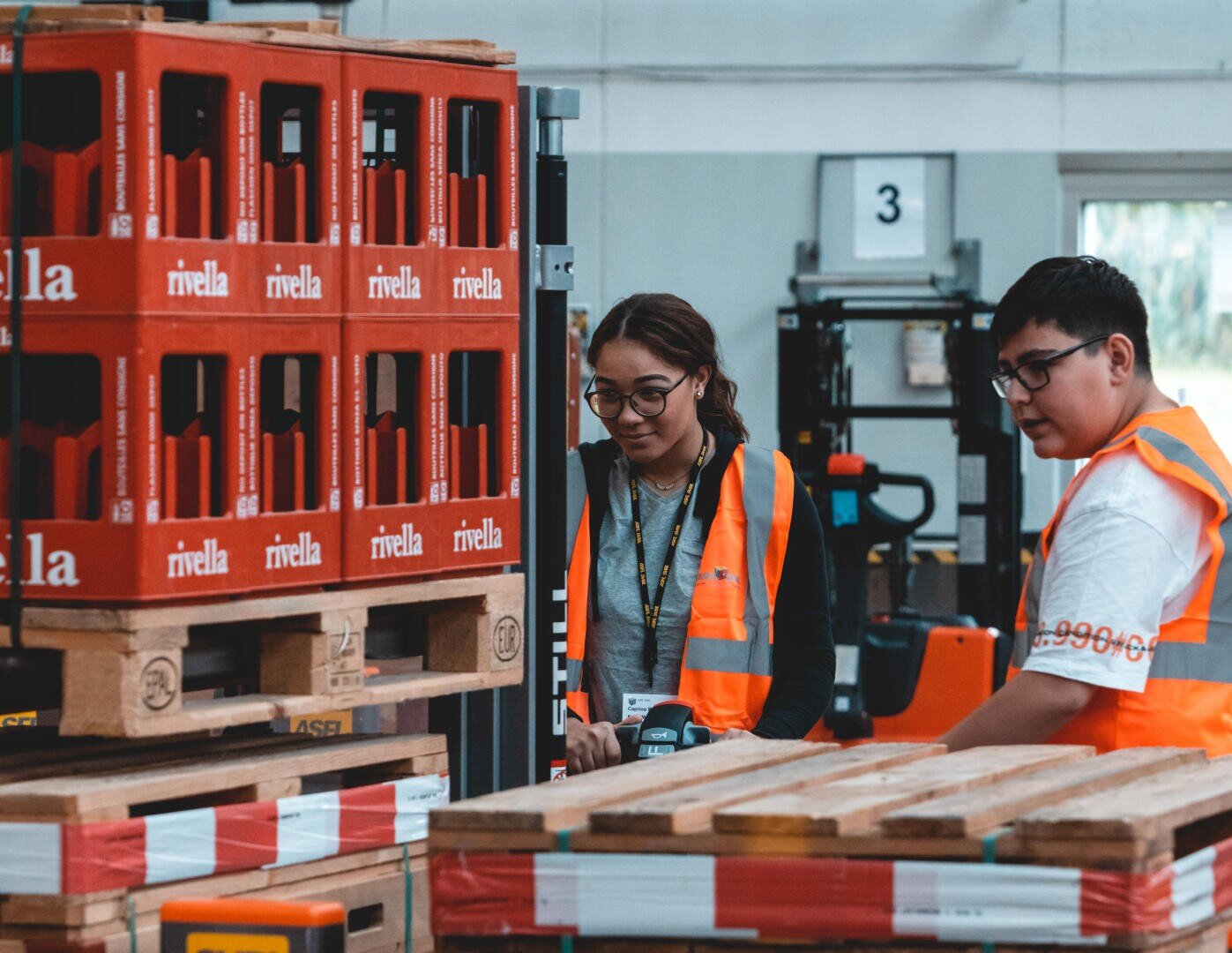 Zwei Personen mit orangefarbenen Sicherheitswesten arbeiten zusammen in einem Lagerhaus. Sie stehen neben Holzpaletten und gestapelten roten Kisten mit der Aufschrift rivella. Im Hintergrund sind Industrieanlagen zu sehen.