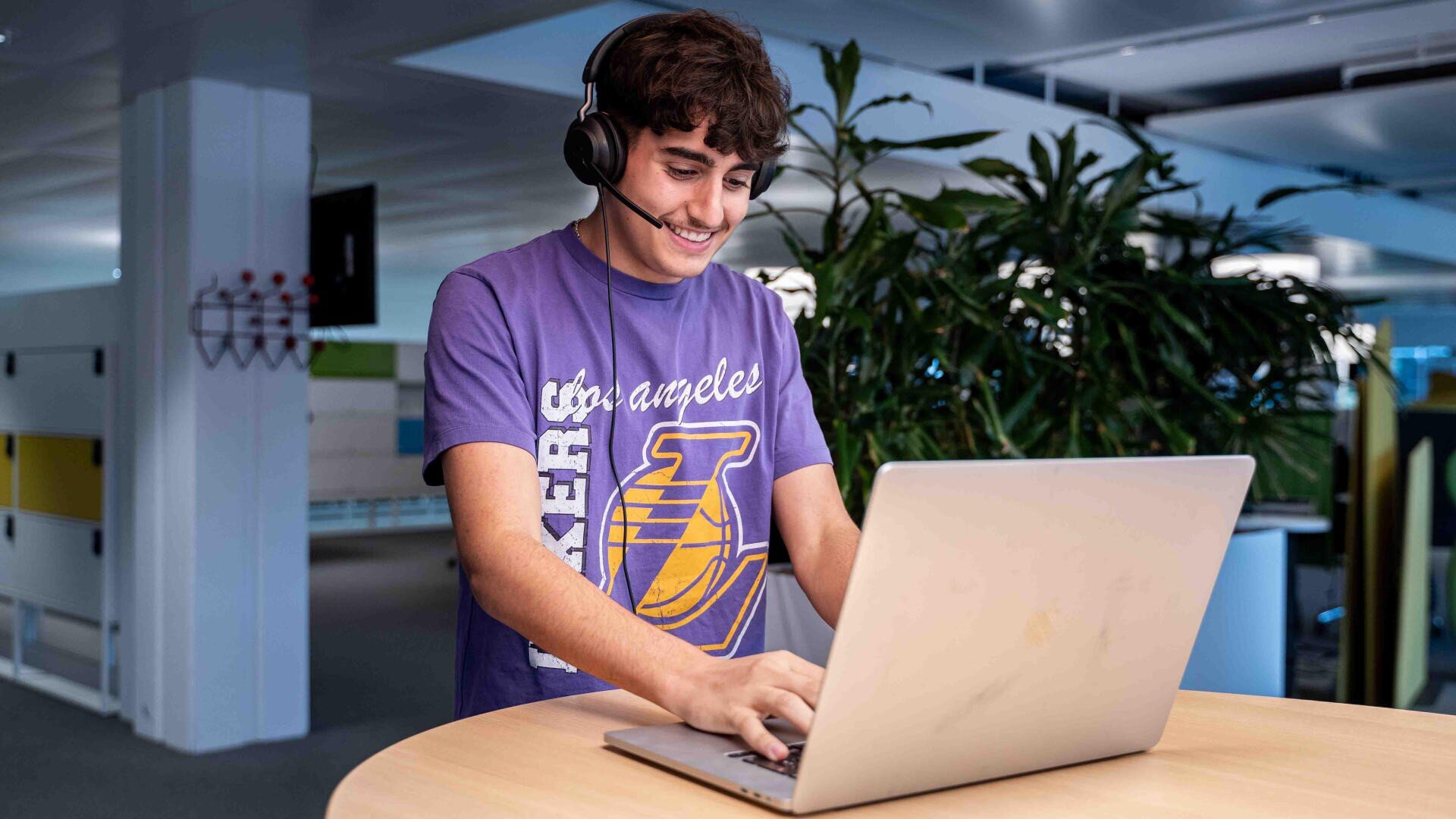 Ein junger Mensch mit Kopfhörern und einem lila Los Angeles Lakers-T-Shirt lächelt, während er an einem runden Tisch in einem modernen, gut beleuchteten Büroraum mit Pflanzen im Hintergrund einen Laptop benutzt.