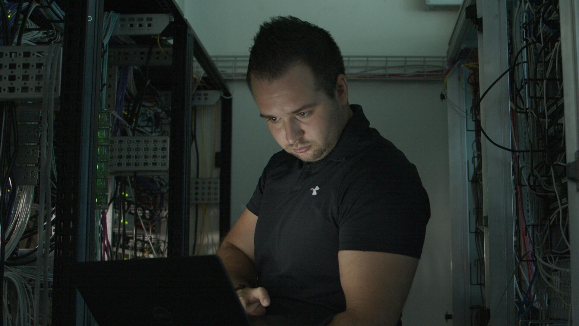 Eine Person in einem schwarzen Hemd arbeitet an einem Laptop in einem schwach beleuchteten Serverraum, umgeben von Netzwerkkabeln und Serverschränken.