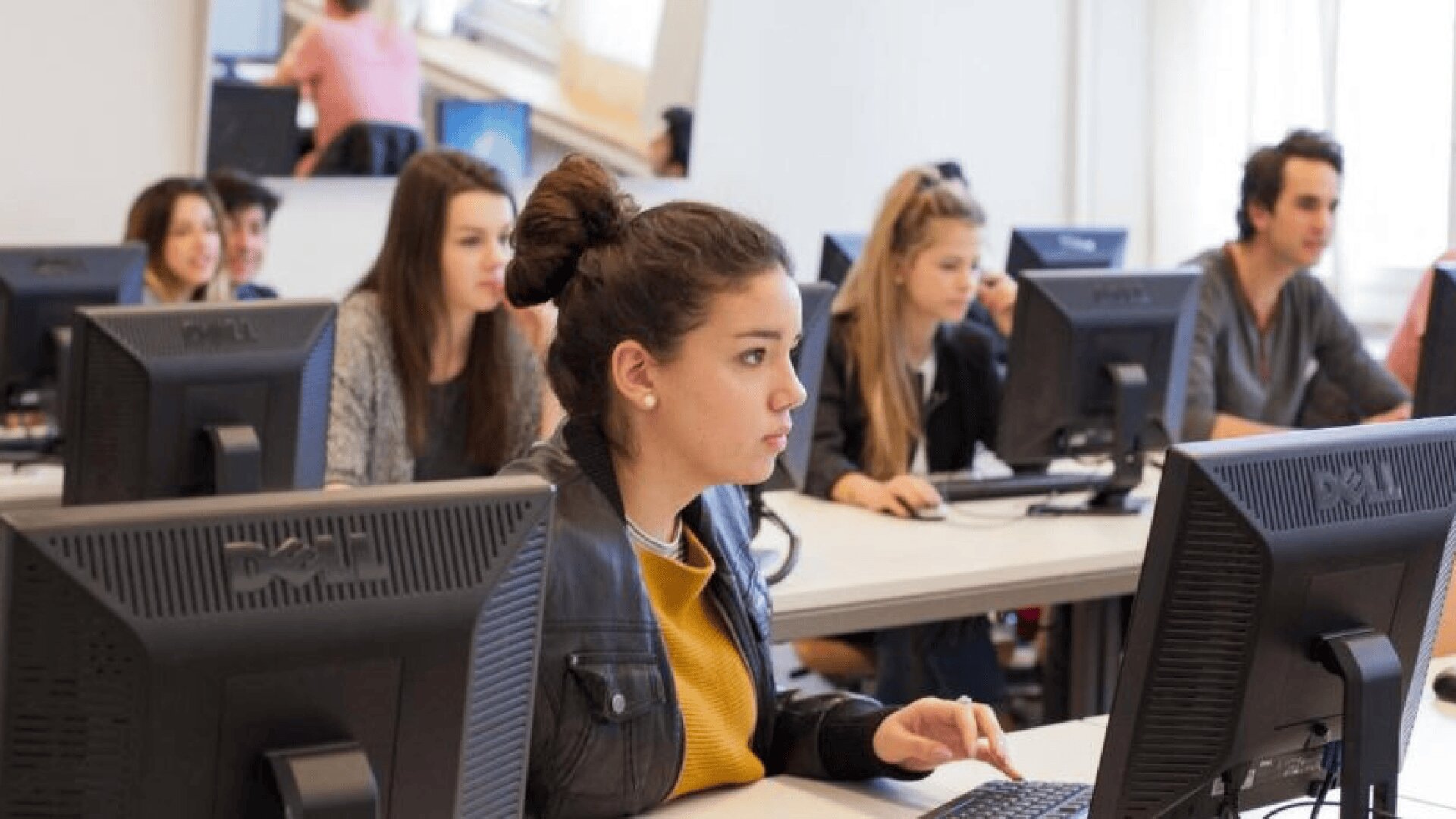 Eine Gruppe von Schülern sitzt an Schreibtischen mit Desktop-Computern in einem hellen Klassenzimmer. Eine junge Frau im Vordergrund konzentriert sich auf ihren Bildschirm, während andere arbeiten oder nach vorne schauen.
