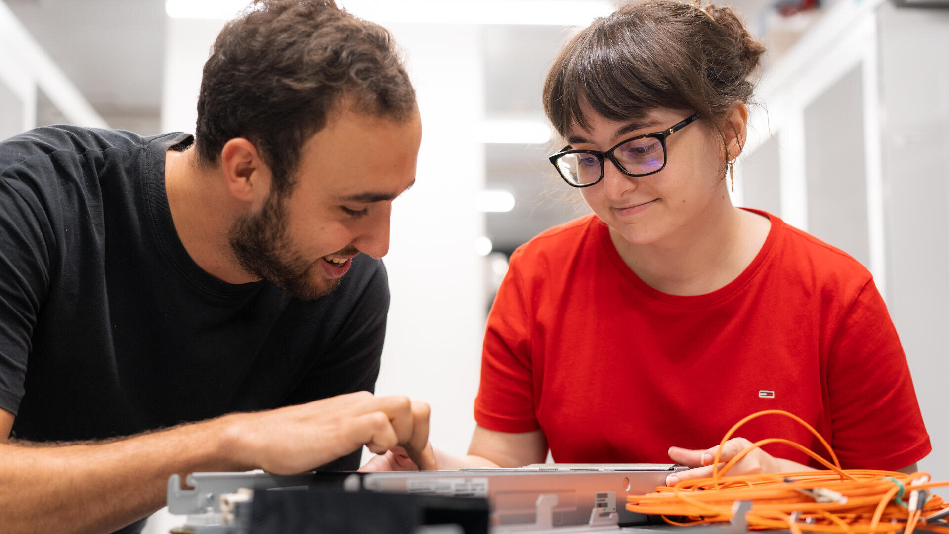 Zwei Personen arbeiten zusammen und untersuchen elektronische Geräte und orangefarbene Kabel auf einem Schreibtisch. Der Mann zeigt auf ein Teil, während die Frau mit Brille und rotem Hemd konzentriert und engagiert wirkt.