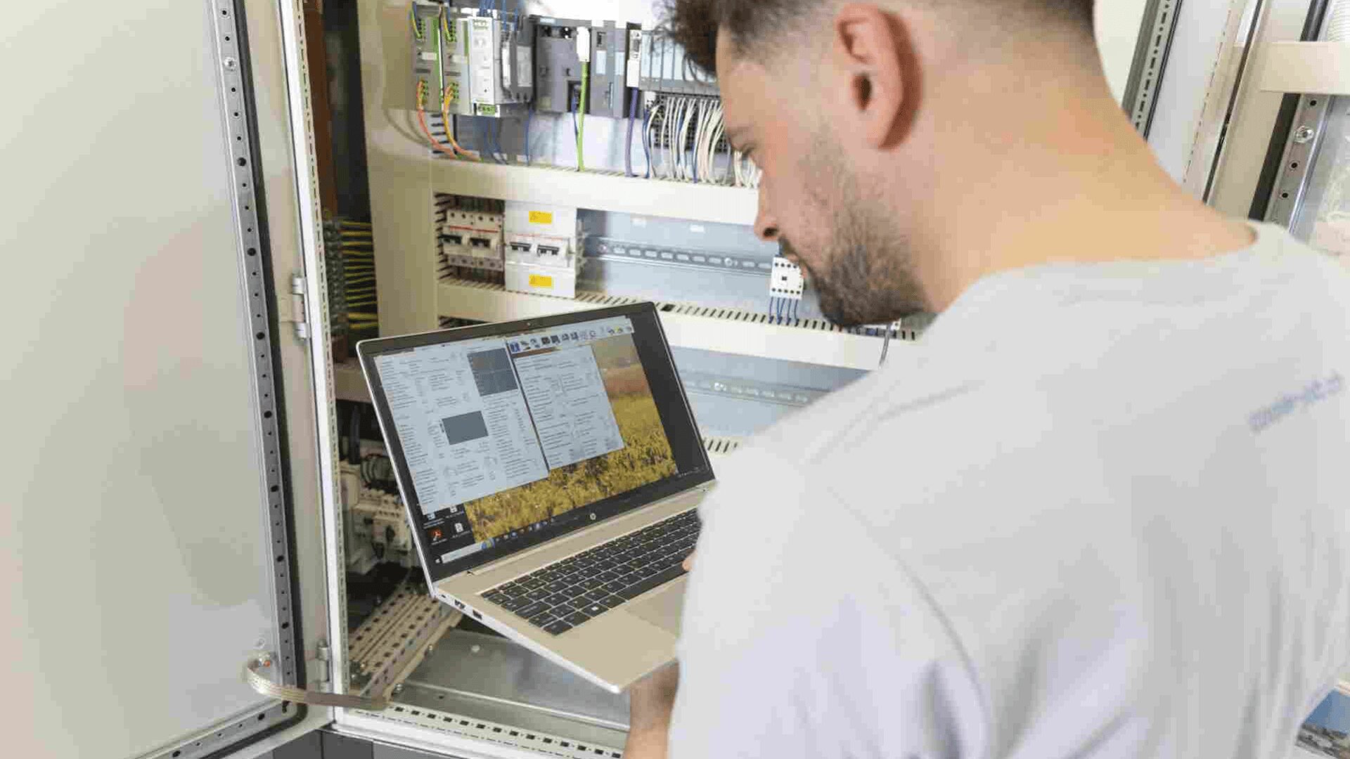 Eine Person steht vor einer offenen elektrischen Schalttafel und hält einen Laptop, auf dem technische Diagramme und Software angezeigt werden. Die Person scheint Geräte zu überwachen oder zu konfigurieren.