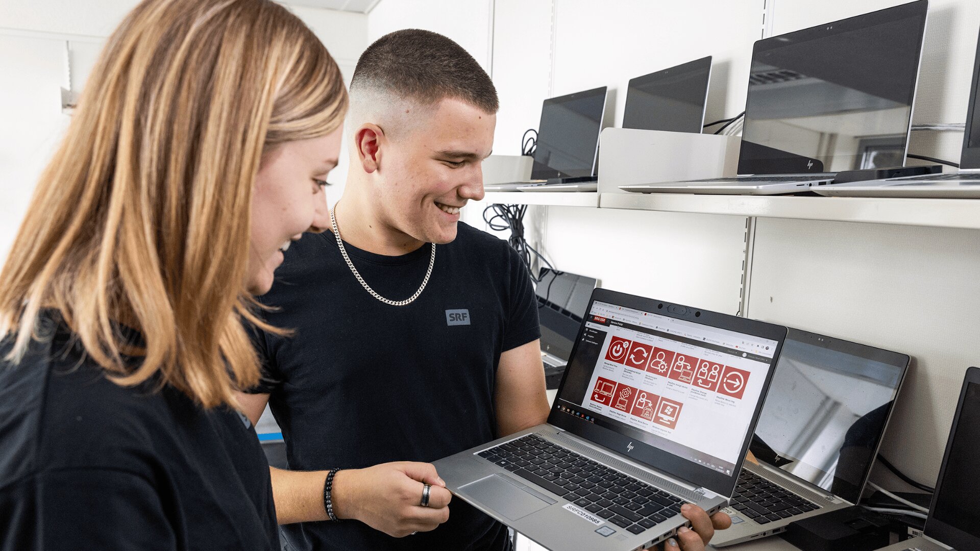 Zwei Personen stehen vor einem Regal mit Laptops. Eine Person hält einen Laptop, der einen roten Bildschirm mit weißen Symbolen zeigt, während beide lächeln und auf den Bildschirm schauen. Im Hintergrund sind weitere Laptops in Regalen zu sehen.