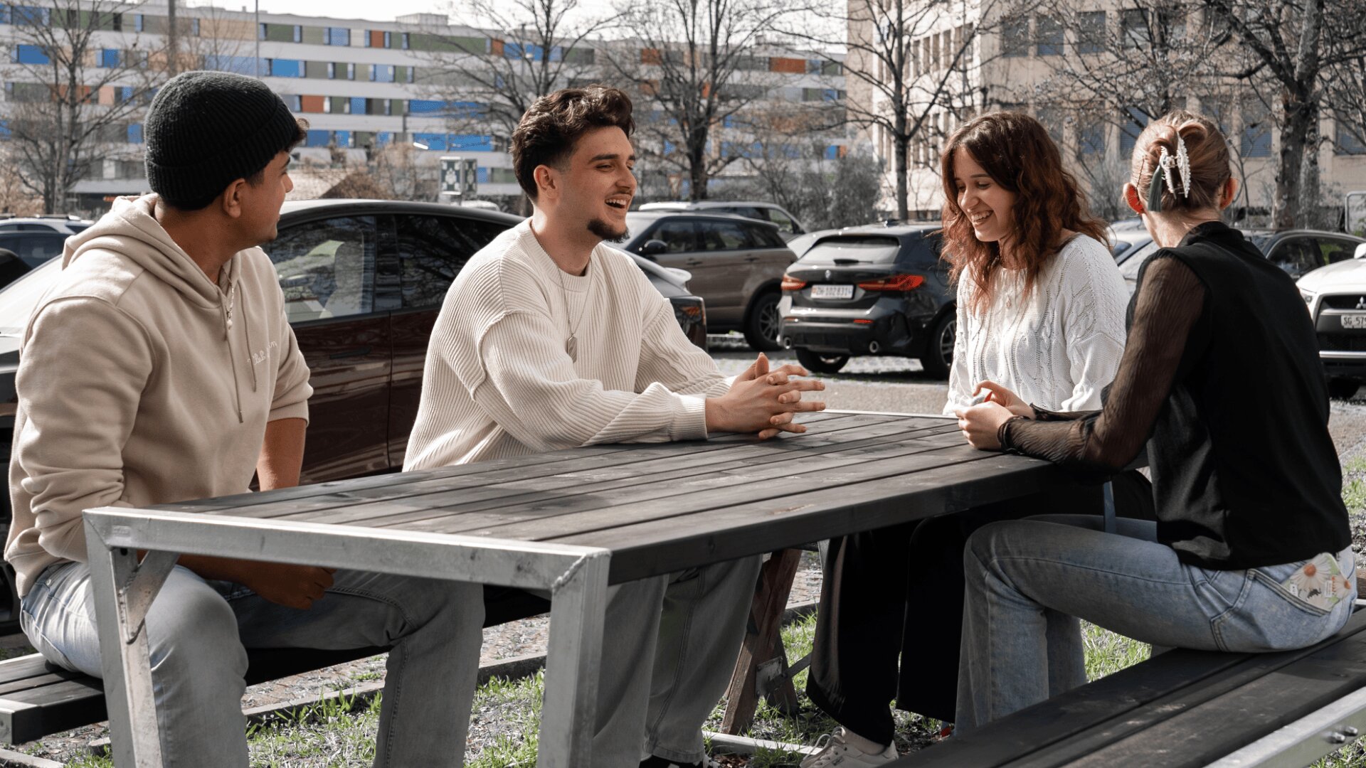 Vier junge Erwachsene sitzen an einem Picknicktisch im Freien, lächeln und reden miteinander. Im Hintergrund sind geparkte Autos und Gebäude zu sehen, und die Gruppe wirkt entspannt und in ein Gespräch vertieft.