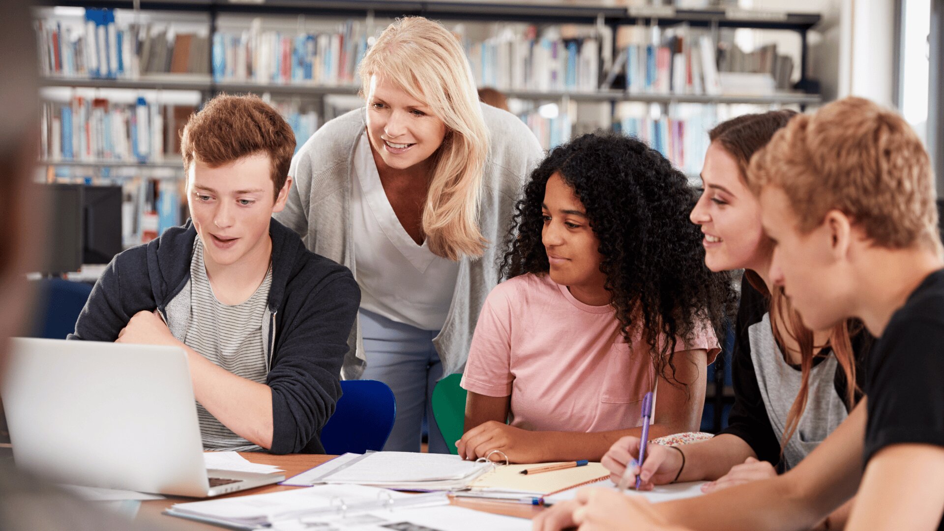 Eine Lehrerin steht und lächelt, während sie vier Highschool-Schülern hilft, die um einen Laptop und Notebooks versammelt sind und in einer Bibliothek zusammenarbeiten. Im Hintergrund sind Bücherregale zu sehen.