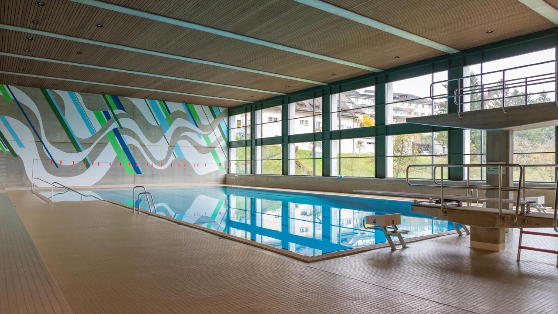 Hallenbad mit klarem Wasser, Sprungbrettern und einem modernen Wandgemälde an der Wand. Große Fenster lassen natürliches Licht herein und geben den Blick auf Gebäude und Grünflächen frei. Der Raum hat eine holzgetäfelte Decke und einen gefliesten Boden.