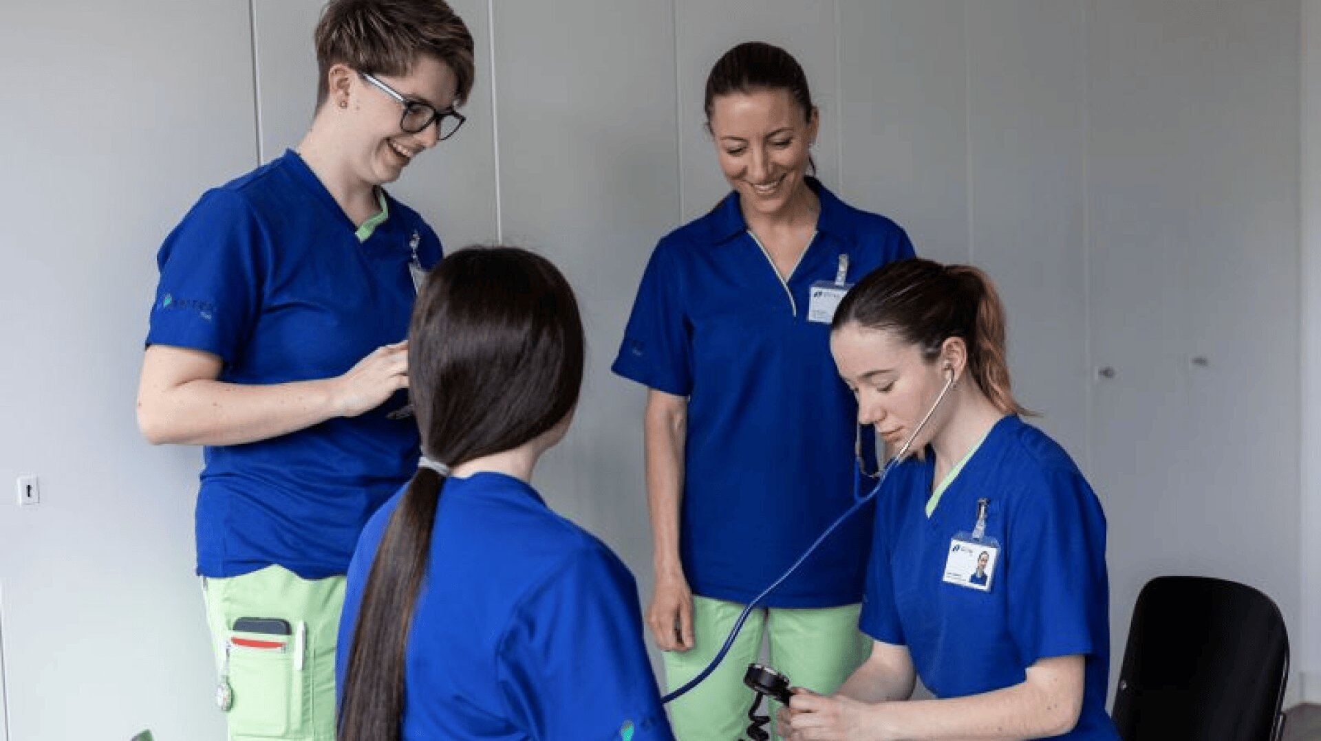 Vier Frauen in blauen Arztkitteln, zwei stehend und lächelnd, eine sitzend mit dem Rücken zur Kamera und eine mit einem Stethoskop, scheinen in einem klinischen Umfeld medizinische Verfahren zu praktizieren.