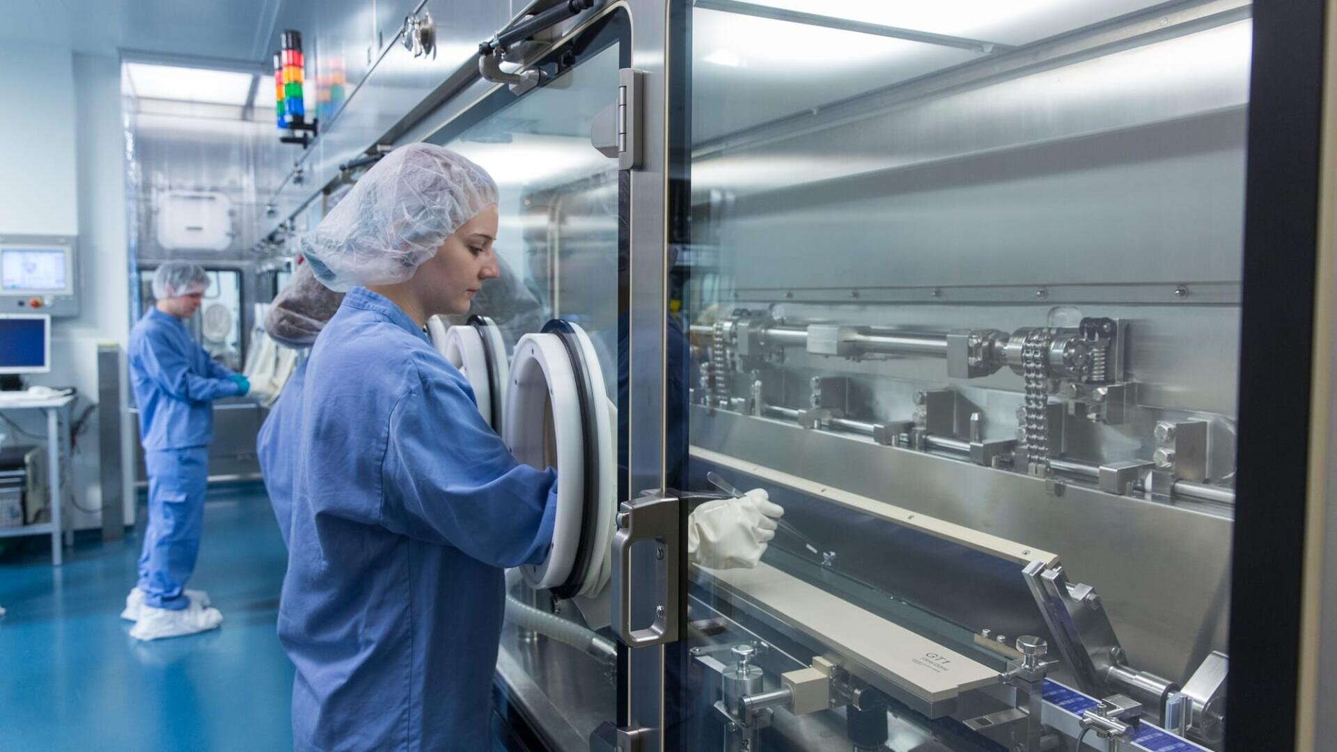 Zwei Wissenschaftler in Schutzkleidung arbeiten in einem sauberen Labor; einer bedient Geräte durch eine versiegelte Glasabdeckung, während der andere im Hintergrund einen Computer bedient.