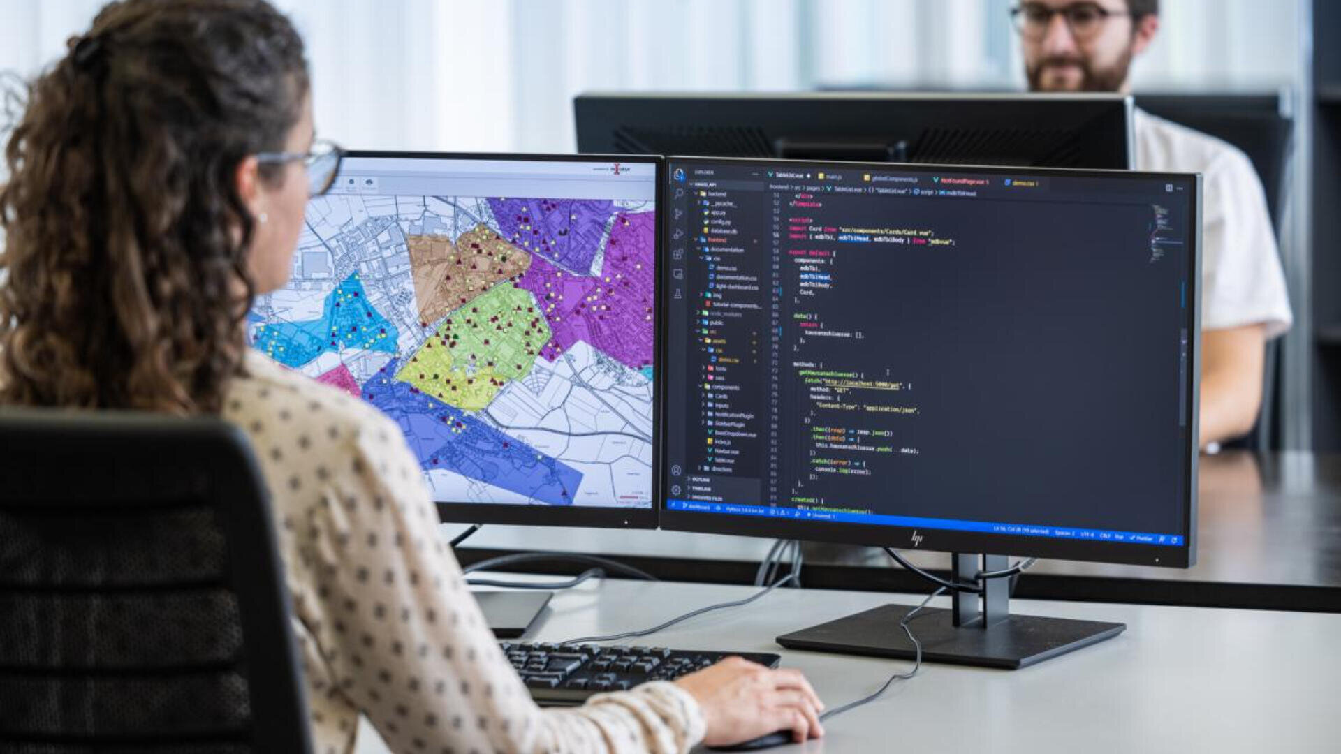 Eine Frau sitzt an einem Schreibtisch mit zwei Monitoren; einer zeigt eine bunte Karte mit Datenpunkten, der andere einen Code. Im Hintergrund arbeitet eine weitere Person an einem Computer.