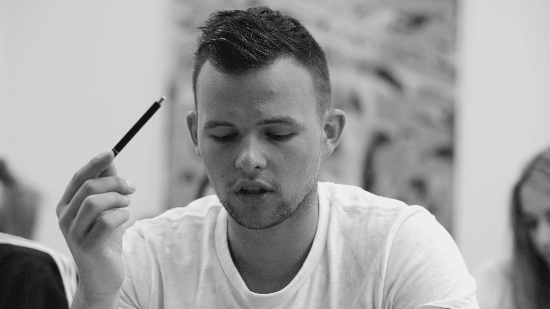 Ein junger Mann in einem weißen Hemd hält einen Stift in der rechten Hand und blickt nachdenklich nach unten, während andere Personen im Hintergrund verschwommen sind. Das Bild ist in schwarz-weiß gehalten.