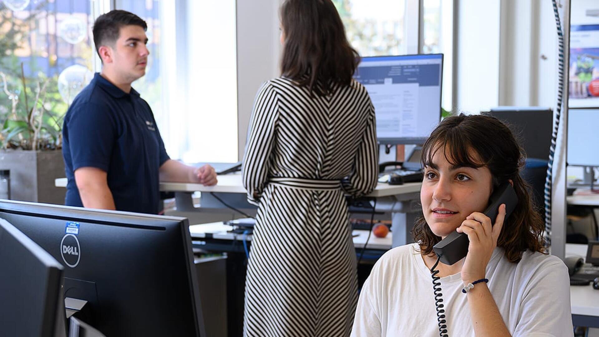 Eine Frau sitzt an einem Schreibtisch und telefoniert mit einem leichten Lächeln. Hinter ihr stehen ein Mann und eine Frau an einem Schreibtisch und unterhalten sich in einem hellen Büro mit Computern und großen Fenstern.