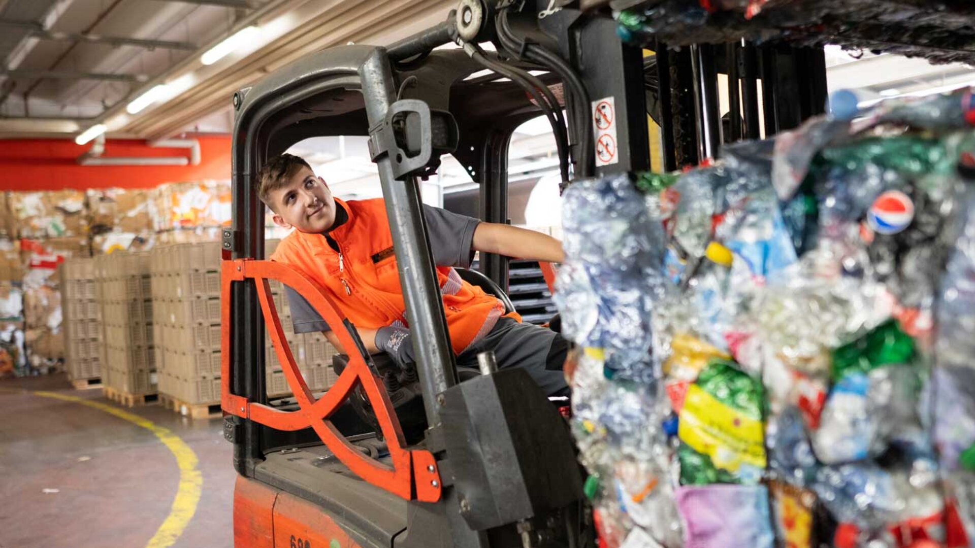 Ein Arbeiter in orangefarbener Uniform bedient einen Gabelstapler, mit dem er gepresste Ballen von Plastikflaschen in einer Recyclinganlage transportiert.