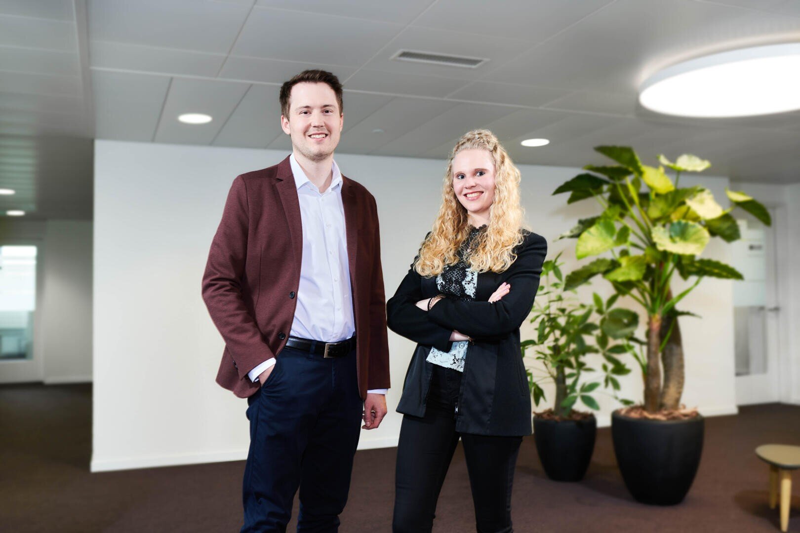 Zwei Fachleute, ein Mann in einem kastanienbraunen Blazer und eine Frau in einem schwarzen Blazer, stehen lächelnd in einem modernen Büroraum mit weißen Wänden, dunklem Teppich und großen Topfpflanzen im Hintergrund.