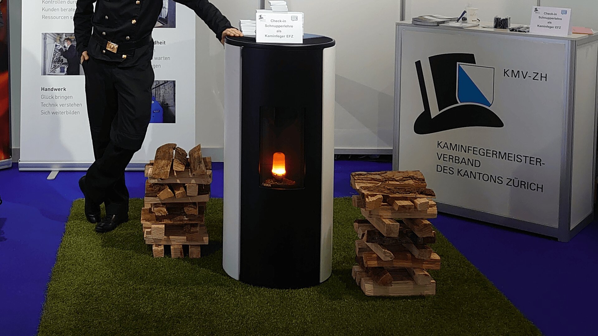 Eine Person in schwarzer Uniform steht neben einem zylindrischen schwarz-weißen Ofen mit einer sichtbaren Flamme im Inneren, umgeben von Stapeln von Brennholz auf Kunstrasen an einem Ausstellungsstand.