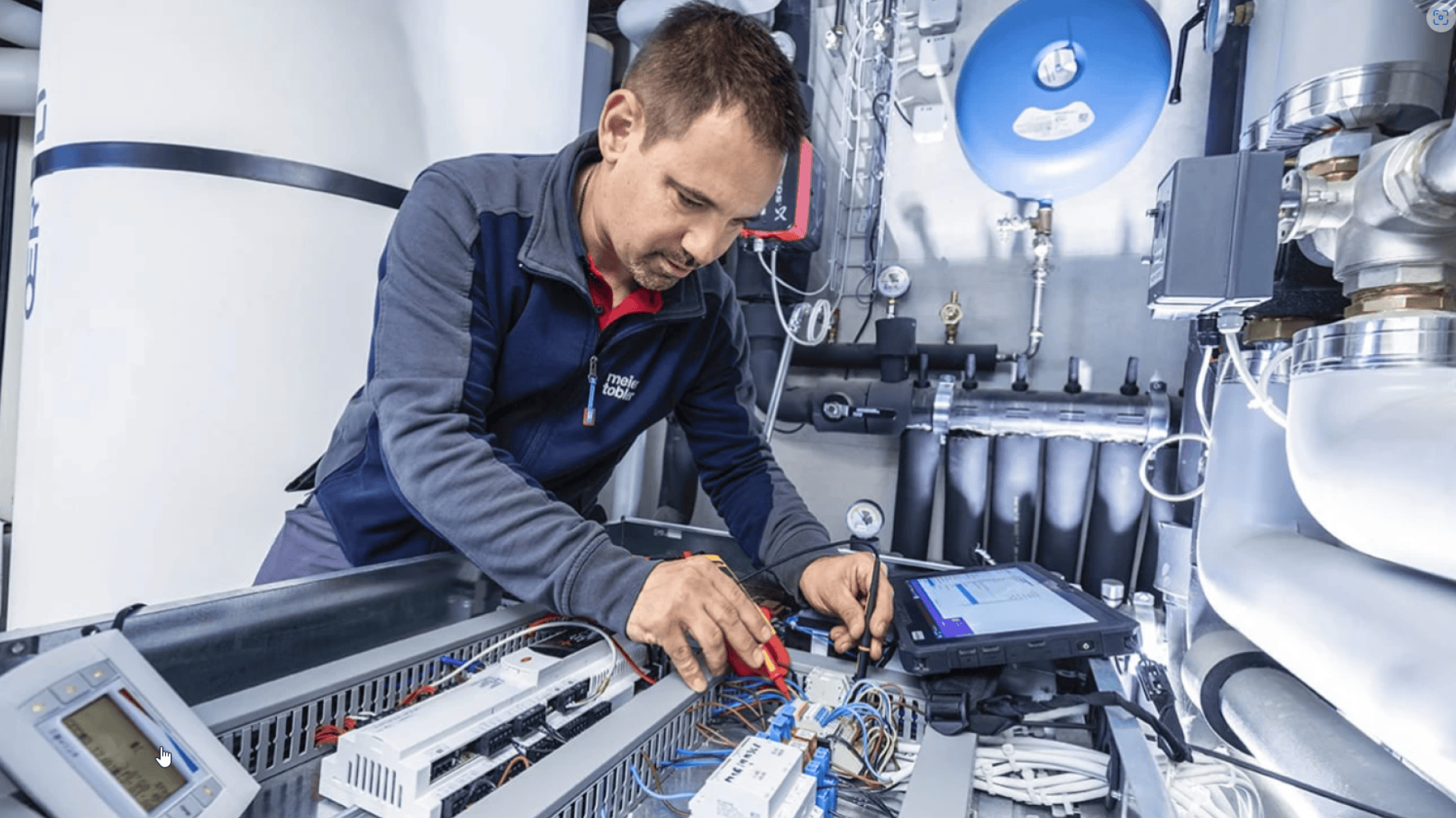 Ein Techniker in blauer Uniform arbeitet mit Werkzeugen an einer offenen Schalttafel voller Drähte und elektronischer Komponenten in einer technischen oder industriellen Umgebung, wobei im Hintergrund verschiedene Geräte und Anlagen zu sehen sind.