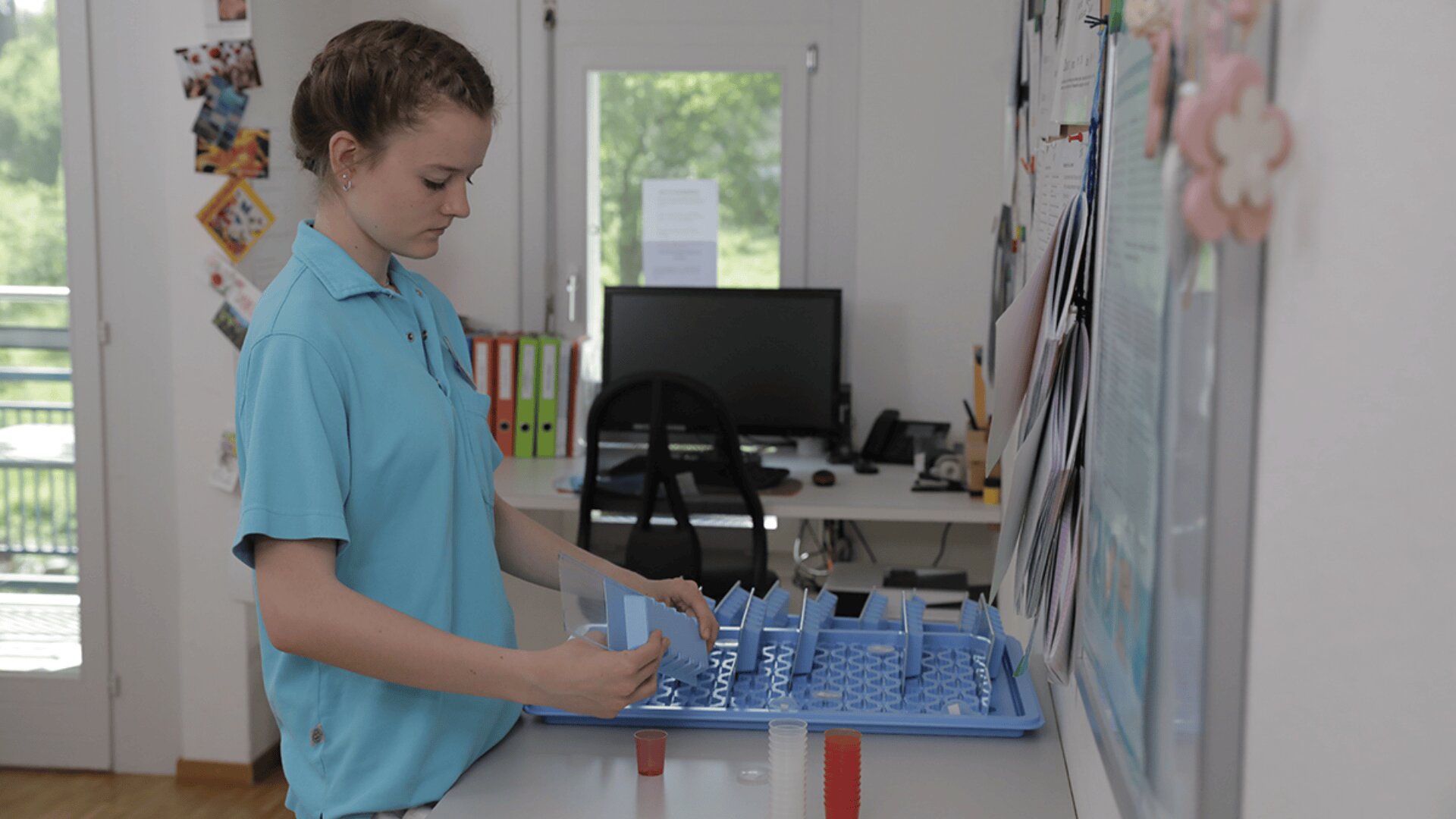 Eine junge Frau in einem blauen Hemd sortiert an einem Schreibtisch in einem gut beleuchteten Büro mit einem Computer, Papierkram und großen Fenstern im Hintergrund Medikamente in einen Pillenorganisator ein.