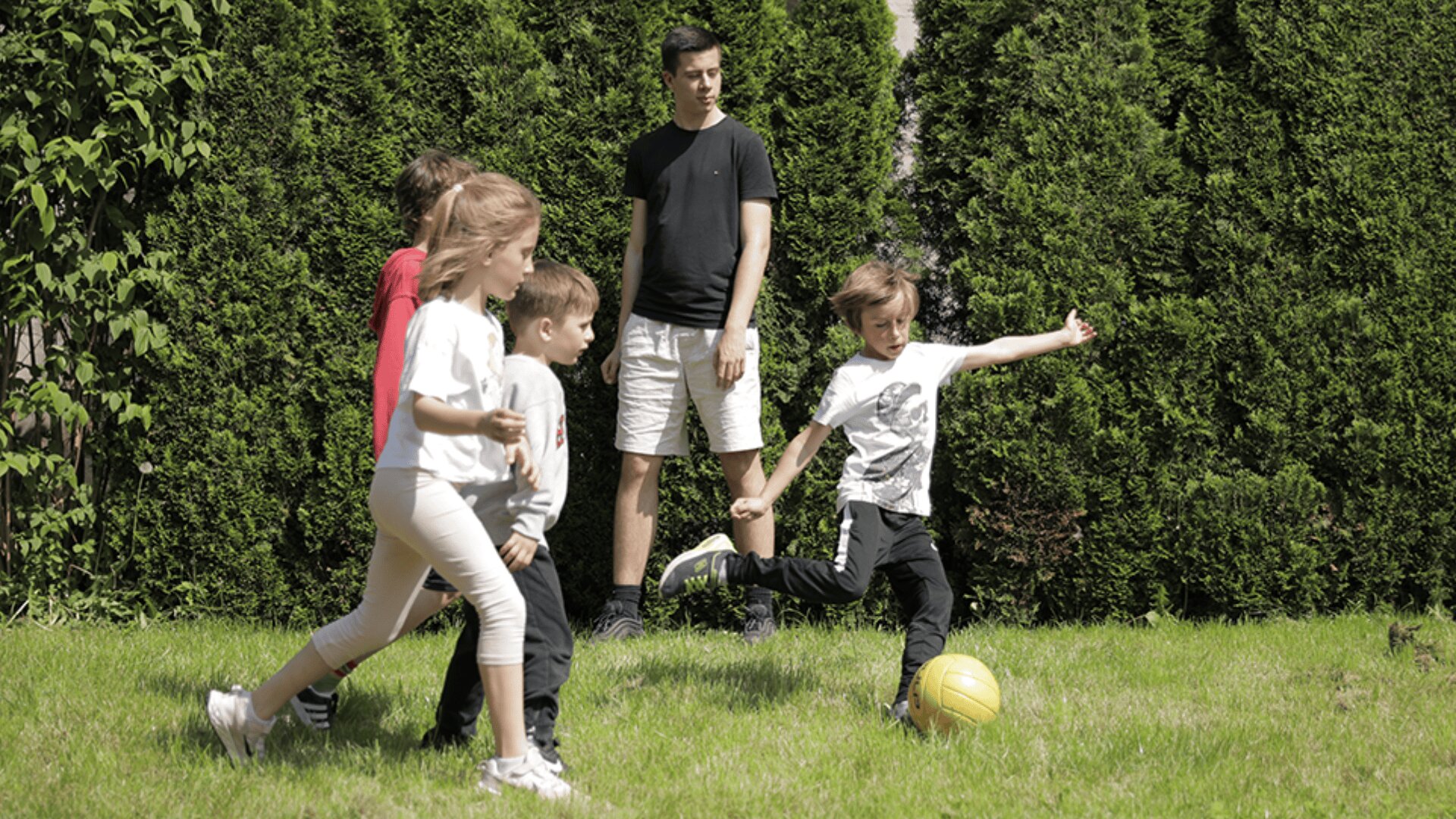 Vier Kinder spielen auf einer Wiese in der Nähe hoher grüner Büsche Fußball, während ein Erwachsener hinter ihnen steht und zusieht. Ein Kind kickt einen gelben Ball, während die anderen in der Nähe laufen.