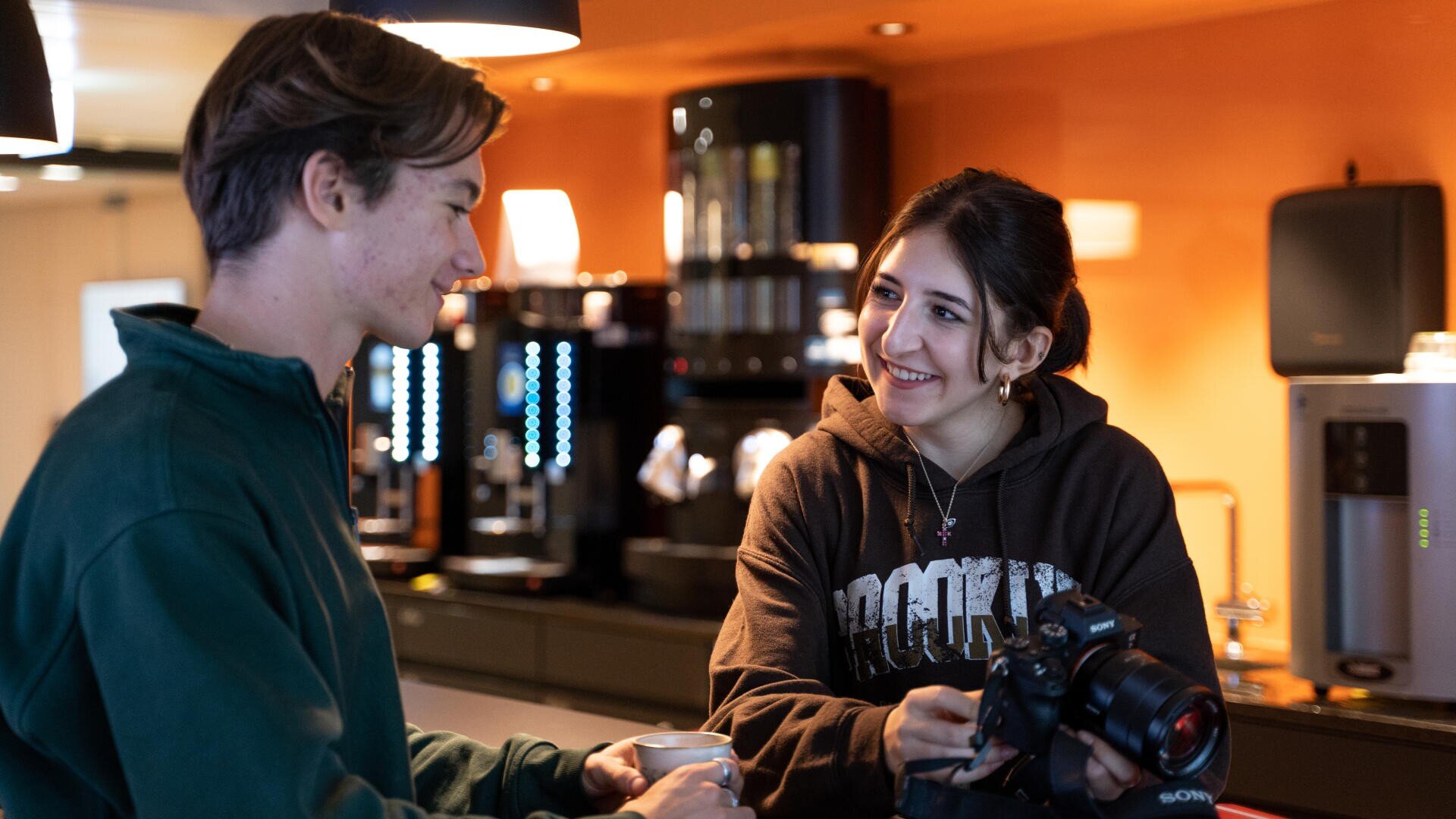 Zwei junge Erwachsene stehen in einem warm beleuchteten Café und unterhalten sich. Der eine hält eine Tasse, der andere eine Kamera und lächelt. Im Hintergrund sind Kaffeemaschinen und eine Arbeitsplatte zu sehen.