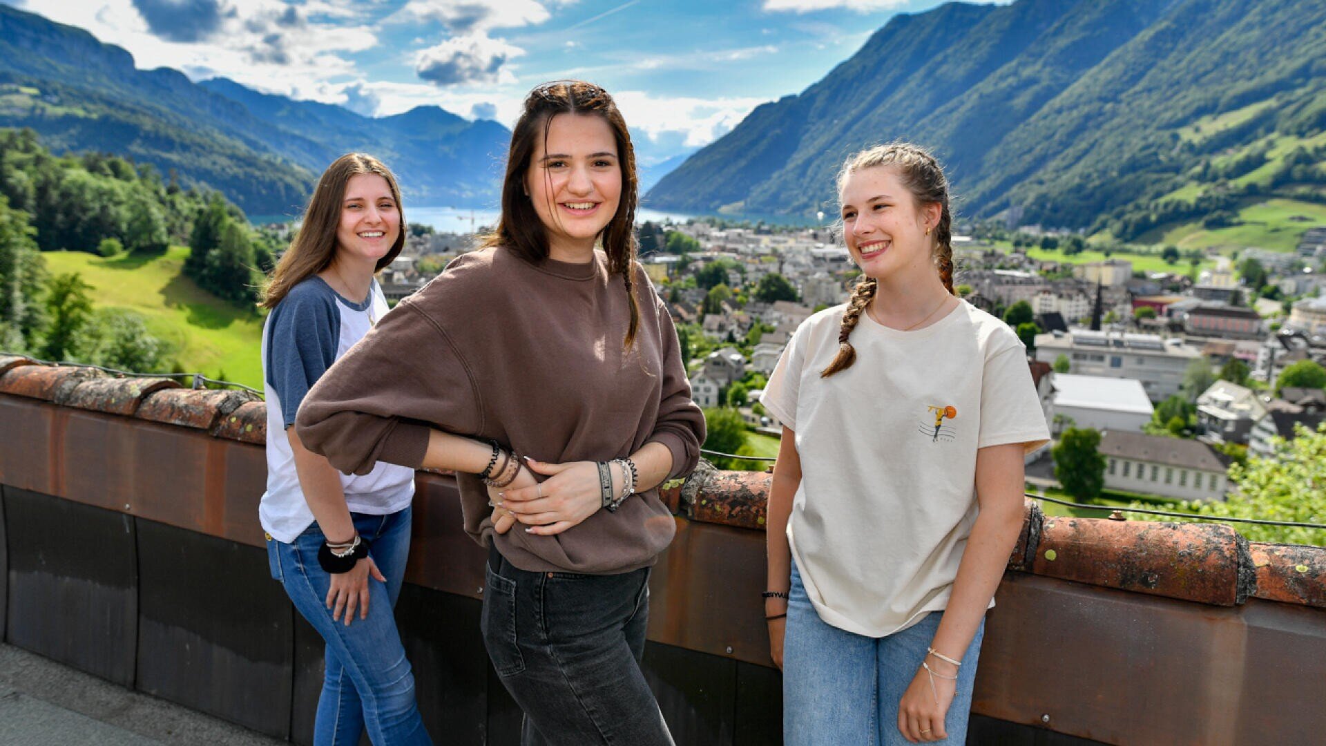 Drei lächelnde Teenager-Mädchen stehen an einem sonnigen Tag im Freien auf einer Terrasse mit malerischen Bergen, einem See und einer Stadt im Hintergrund.