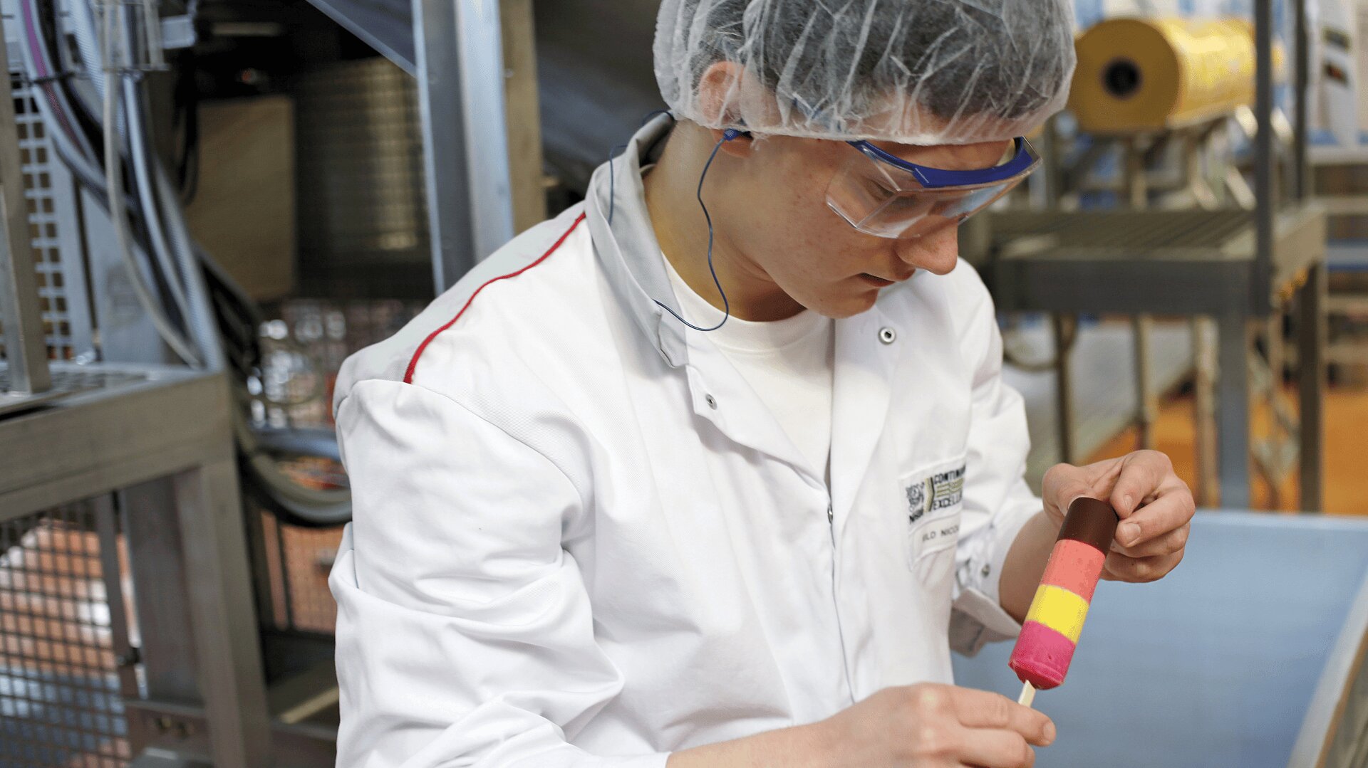 Eine Person in weißem Laborkittel, Haarnetz und Schutzbrille untersucht einen bunten, geschichteten Eisriegel in einer Lebensmittelproduktionsanlage.