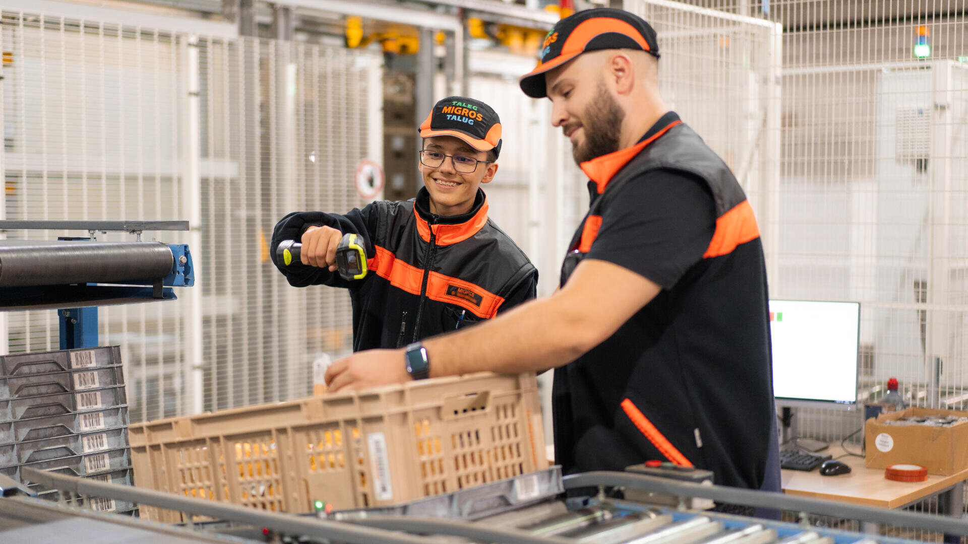 Zwei Arbeiter in schwarz-orangener Uniform bedienen Maschinen in einem Lagerhaus. Einer benutzt ein Elektrowerkzeug, während der andere eine Plastikkiste hält. Im Hintergrund sind Regale, Sicherheitsschranken und ein Computer zu sehen.