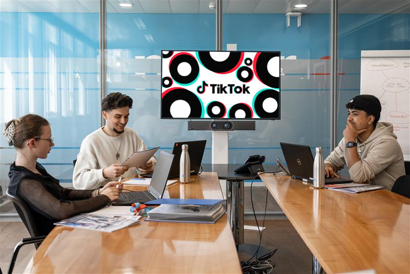 Drei junge Erwachsene sitzen mit Laptops und Papieren an einem Konferenztisch und halten eine Besprechung ab. Auf einem Bildschirm sind das TikTok-Logo und bunte Kreise im Hintergrund zu sehen. Der Raum hat Glaswände und ein modernes Design.