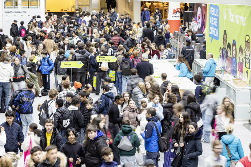 Eine große Menschenmenge füllt ein belebtes Kongresszentrum, in dem die Teilnehmer an den Check-in-Schaltern Schlange stehen. Schilder und ein "Willkommen"-Banner sind zu sehen. Viele Menschen tragen Jacken und Rucksäcke.