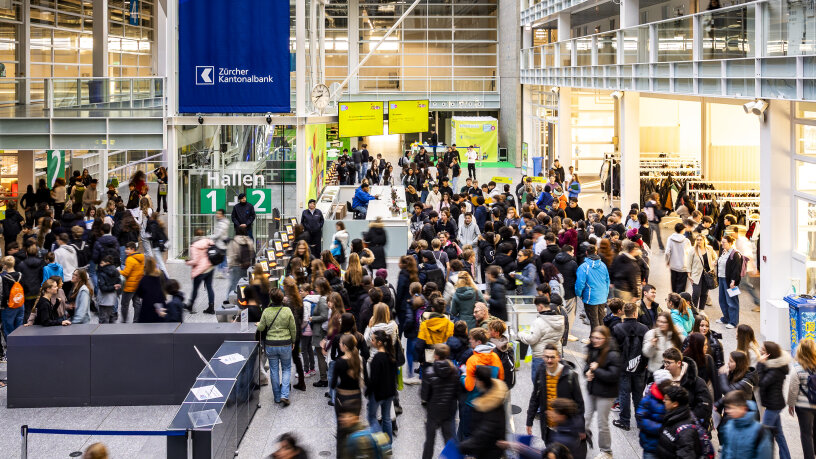 Eine grosse Menschenmenge, viele mit Winterjacken und Rucksäcken, geht durch eine belebte, moderne Halle mit Glaswänden, digitalen Bildschirmen und Schildern mit der Aufschrift "Hallen 1+2" und "Zürcher Kantonalbank".