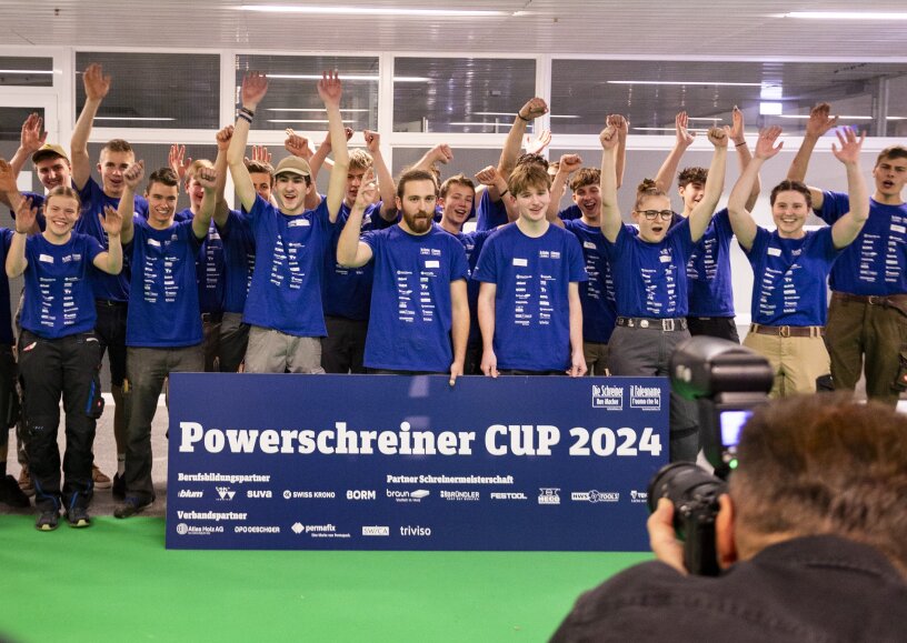 Eine Gruppe junger Leute in blauen Hemden steht jubelnd hinter einem Powerschreiner CUP 2024-Schild, während ein Fotograf den Moment in einer Halle festhält.