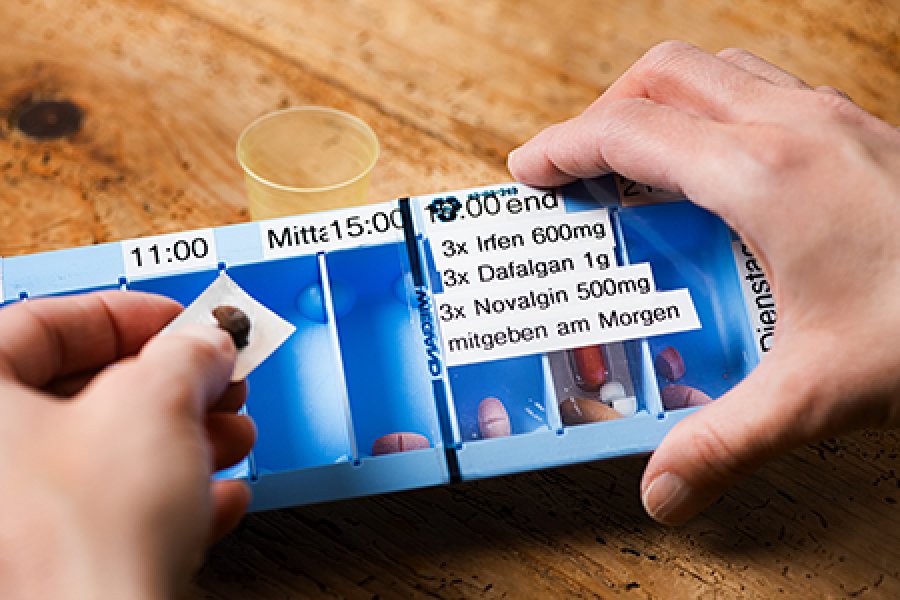 Die Hände einer Person sortieren Pillen in einen blauen Pillenordner, der mit Zeiten und Tagen in deutscher Sprache beschriftet ist; ein Medikamentenbecher und ein Papier mit Medikamentenanweisungen sind auf einem Holztisch zu sehen.