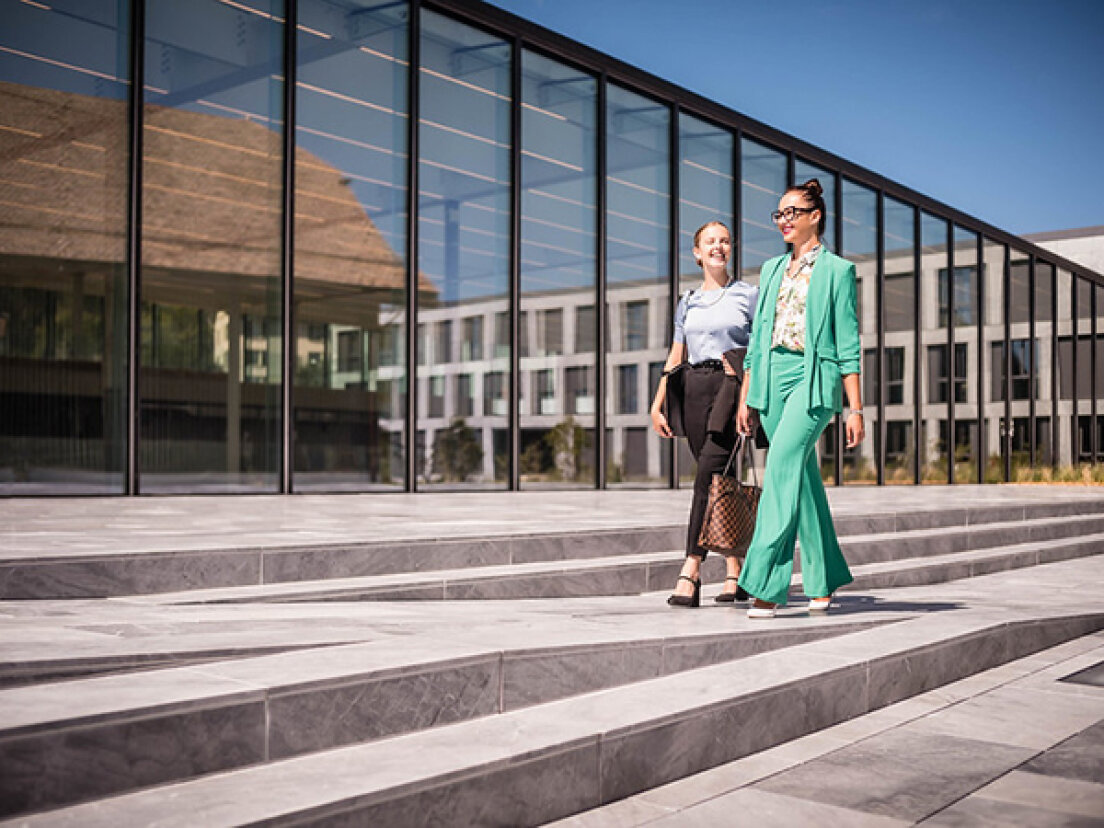 Zwei Frauen in Geschäftskleidung gehen nebeneinander auf breiten Steinstufen vor einem modernen Bürogebäude aus Glas, lächeln und unterhalten sich an einem sonnigen Tag.