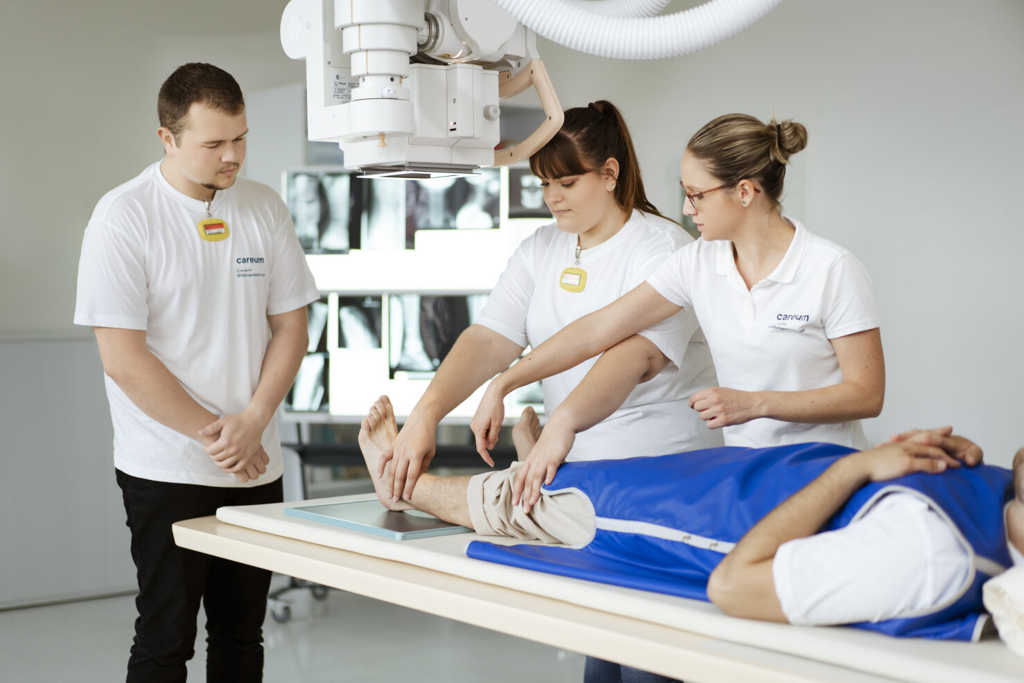 Drei Mediziner in weißen Uniformen positionieren das Bein eines Patienten unter einem Röntgengerät, während eine weitere Person zuschaut. Der Patient liegt auf einem Tisch mit einer blauen Schutzabdeckung. Im Hintergrund sind Röntgenbilder zu sehen.