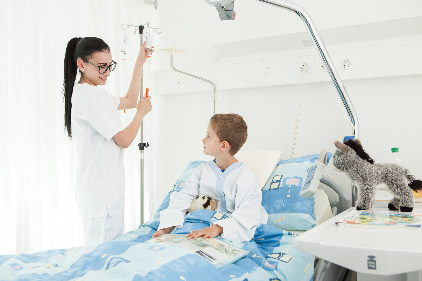 Eine Krankenschwester justiert einen Tropf neben einem lächelnden Jungen, der in einem Krankenhausbett liegt und ein Stofftier hält. Ein weiteres Stofftier liegt auf dem Bett, und das Zimmer wirkt hell und sauber.
