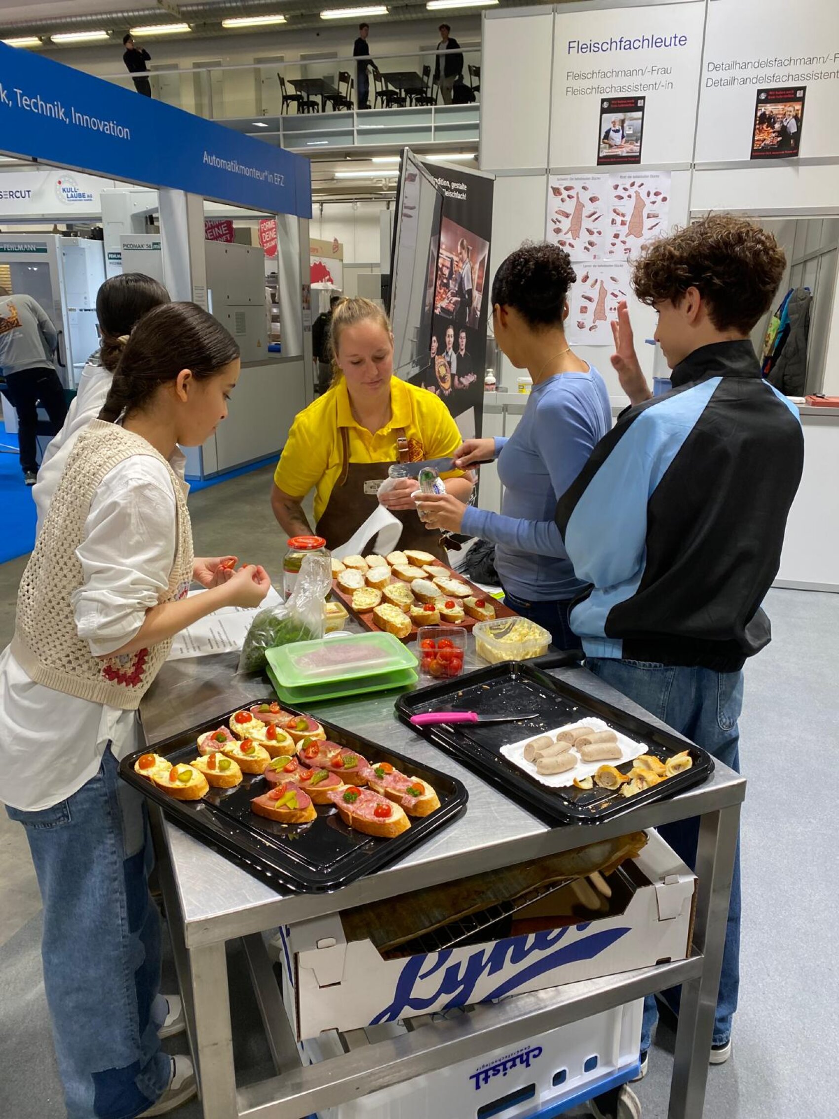 Vier Personen stehen um einen Imbisswagen herum und stellen bei einer Indoor-Veranstaltung Sandwiches mit frischen Belägen zusammen. Im Hintergrund sind Messestände und Banner zu sehen.